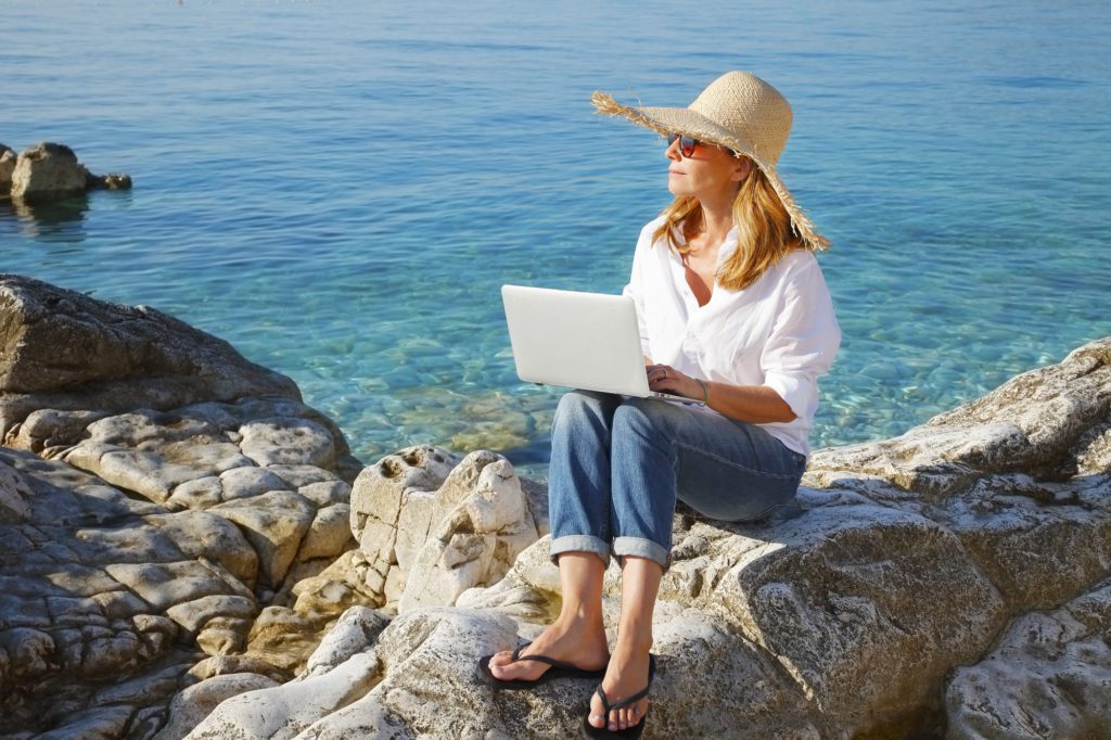 Portrait of beautiful business woman sitting by the sea and working online with her laptop. Middle age female wearing straw hat and sunglasses.